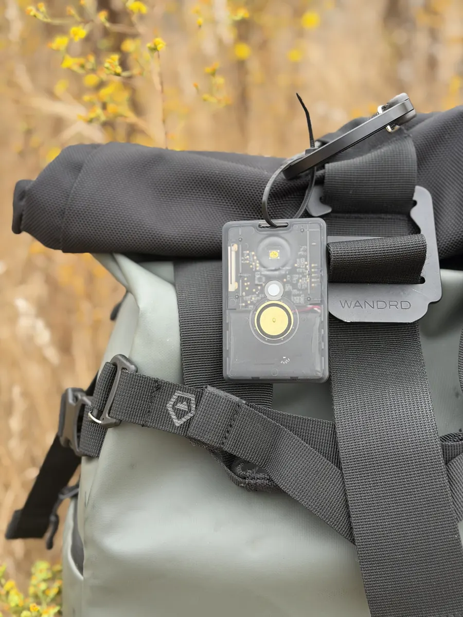 Clear-cased mesh radio test device clipped to a backpack strap, dry chaparral brush in the background