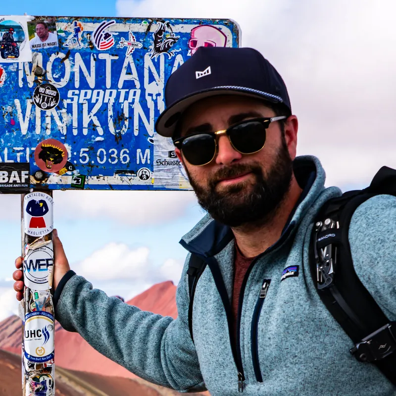 Cameron Cooper, founder of Smoke Signal, in a fleece and cap beside the Vinicunca summit sign